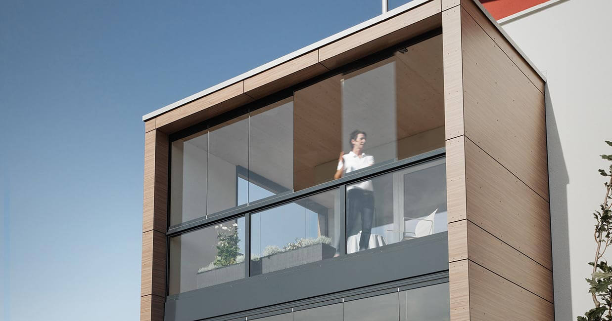 Bild på hus med stora skjutpartier på balkong och en kvinna som står på insidan och tittar ut. 