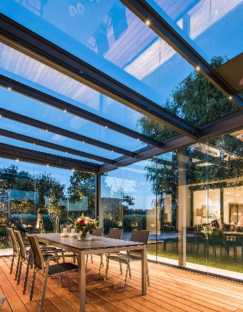 Smuk udestue i glas og aluminium set indefra om aftenen indrettet med spisebord og trægulv.