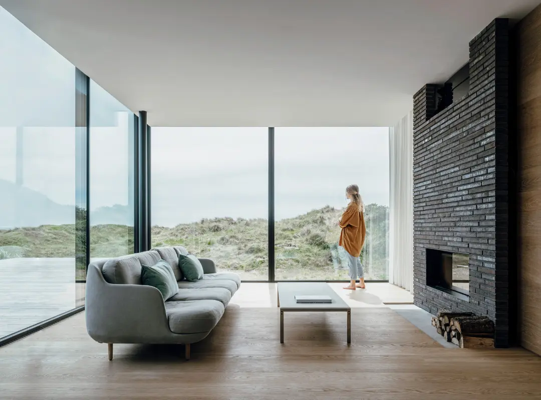 Skydedør med store glaspartier set indefra stue med grå sofa, pejs og flot udsigt ud over strand. Skydedørene giver et syn uden forhindringer ud over stranden.