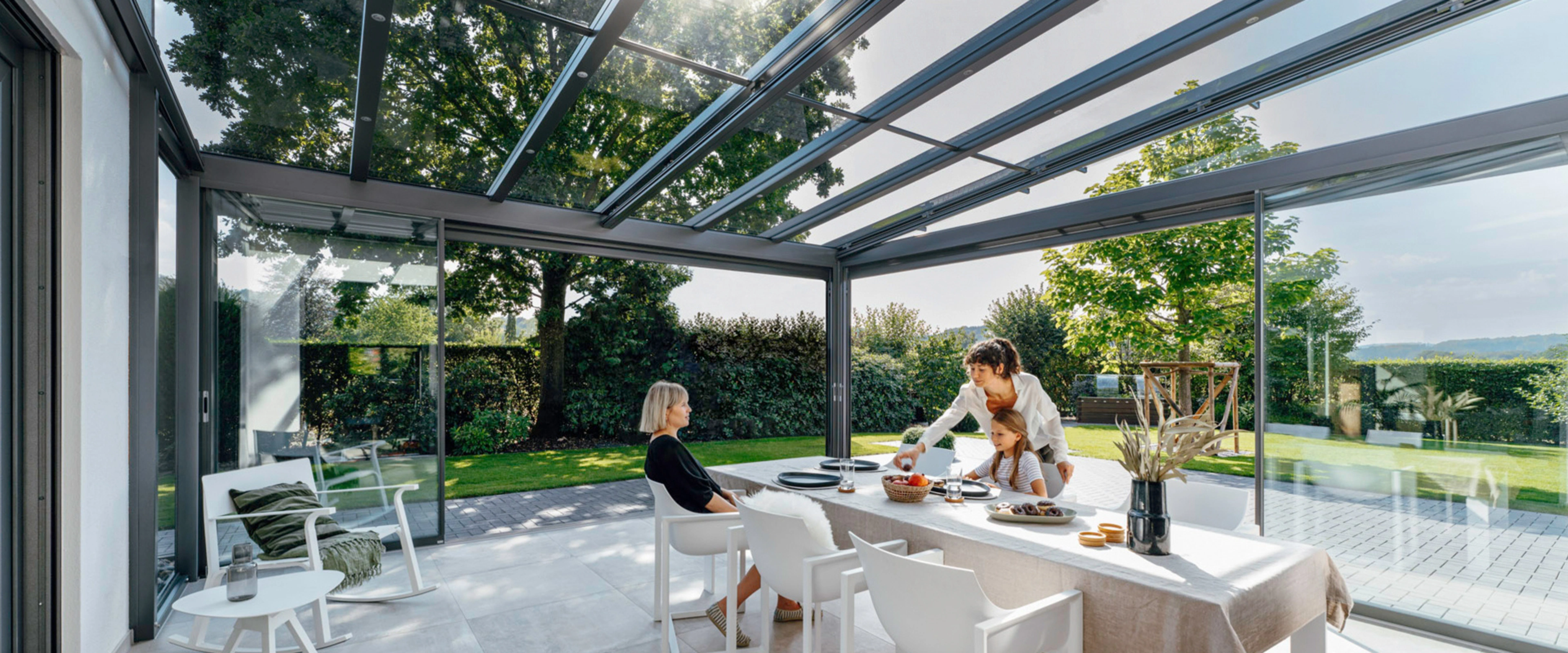To kvinder og en pige sidder ved spisebordet i en glasinddækket terrasse.
