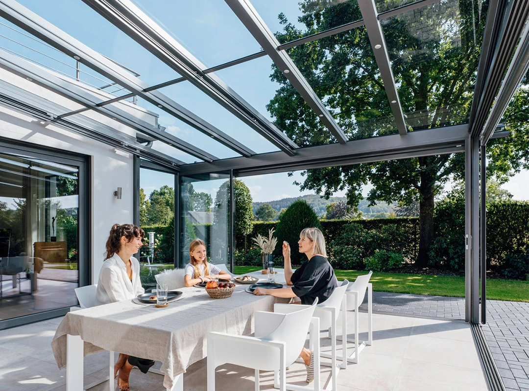 To kvinder og en pige sidder ved spisebordet i en glasinddækket terrasse.
