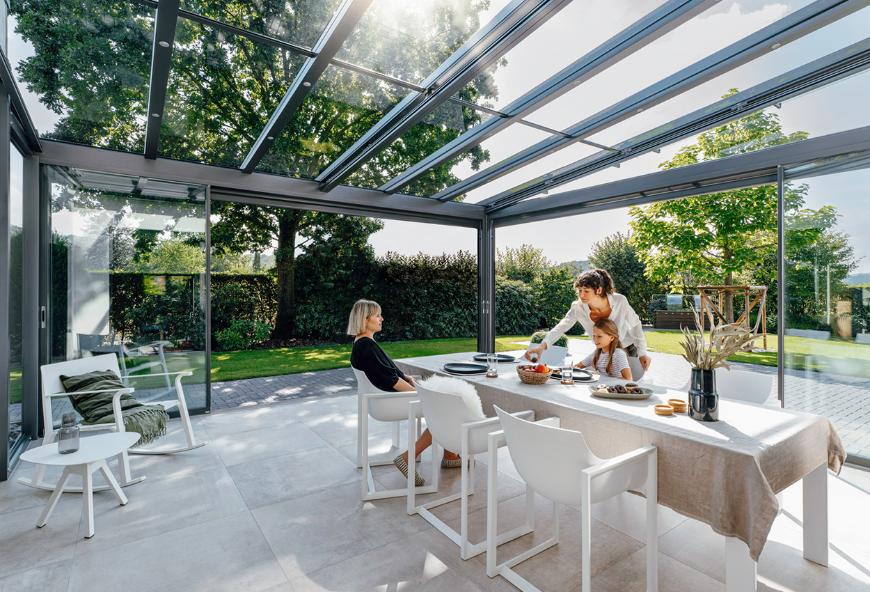 To kvinder og en pige sidder ved spisebordet i en glasinddækket terrasse. 