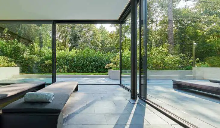 Skydedør med store glaspartier, som set indefra giver adgang til udeareal, som er pool- og terrasseområde. Indenfor er der placeret to daybeds, hvorpå der er lagt et håndklæde.