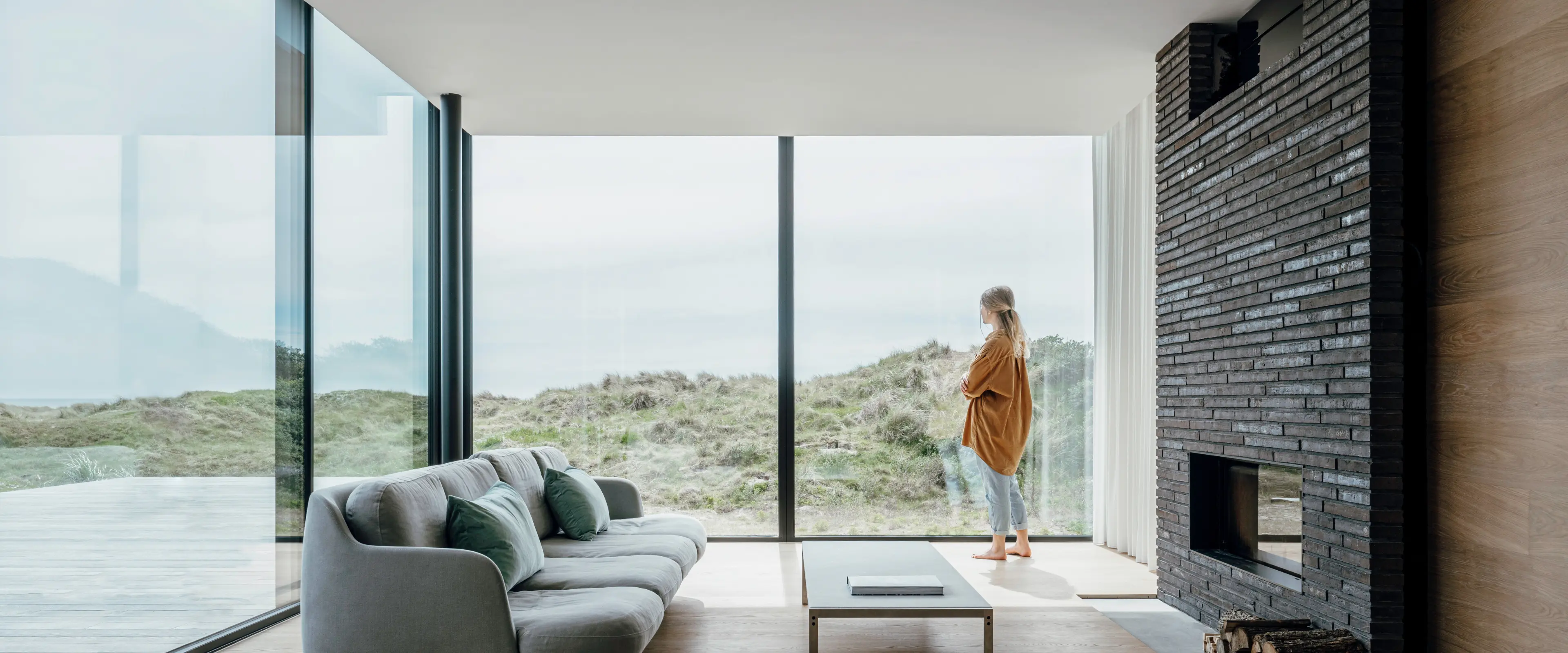 Skjutdörr med stora glaspartier sett inifrån vardagsrummet med grå soffa, öppen spis och fantastisk strandutsikt. Skjutdörrarna ger en fri utsikt över stranden.