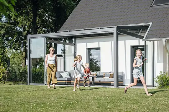 Mutter steht vor einer Glas-Terrasse und schaut Kindern beim Spielen zu